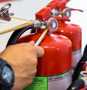 A person conducts a fire inspection, carefully examining a red dry chemical fire extinguisher while holding a pen near the pressure gauge.