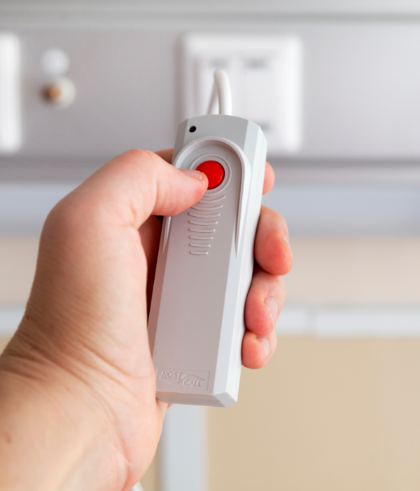 A person holding a white handheld device with a red button, likely part of nurse call systems used for initiating a call or alert, with electrical outlets in the background.