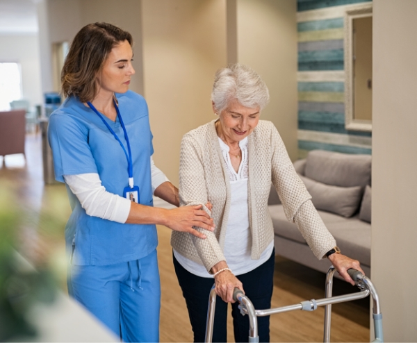 A healthcare worker in blue scrubs assists an elderly woman using a walker in a well-lit, modern assisted living room.