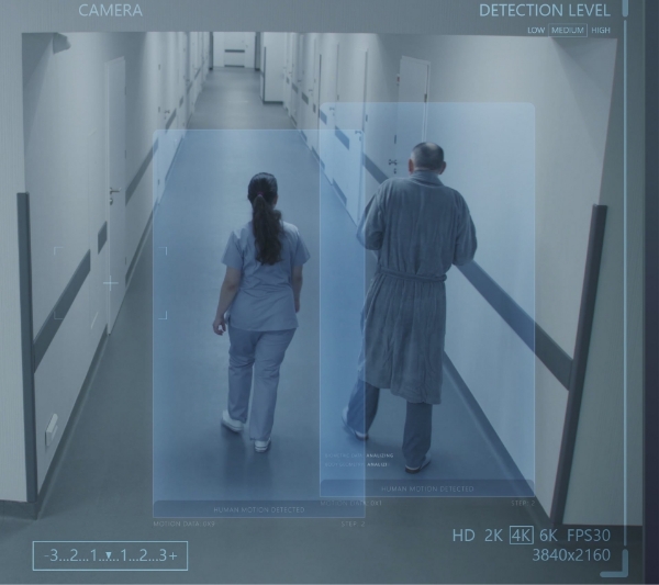 A person in a hospital gown walks down a hallway, accompanied by another individual in scrubs. The scene, viewed through a security camera display, emphasizes the importance of assisted living and fire protection in healthcare settings.