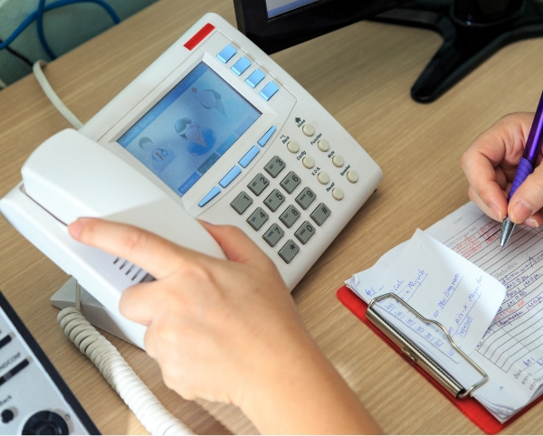 A person holding a phone handset while taking notes on a notepad. On the desk, a white telephone with a digital screen displaying people icons sits nearby, ensuring seamless communication in assisted living.