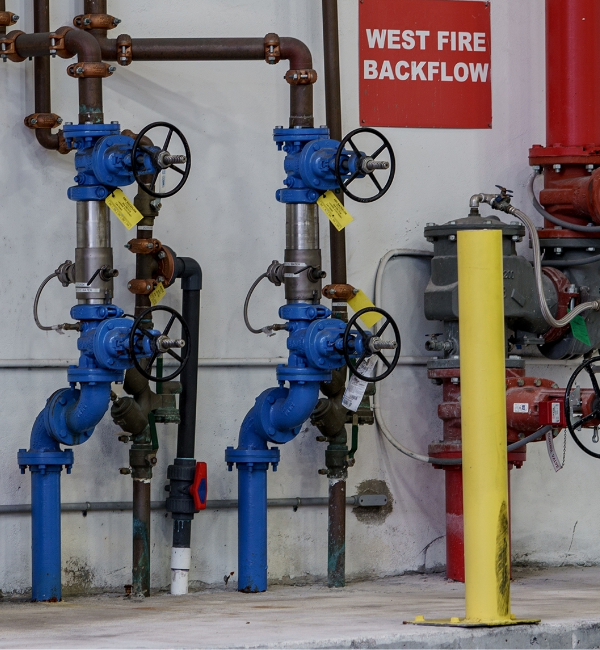 Two blue valves with circular handles, labeled "West Fire Backflow," are installed on pipes in an industrial setting. A yellow pole stands nearby for protection.